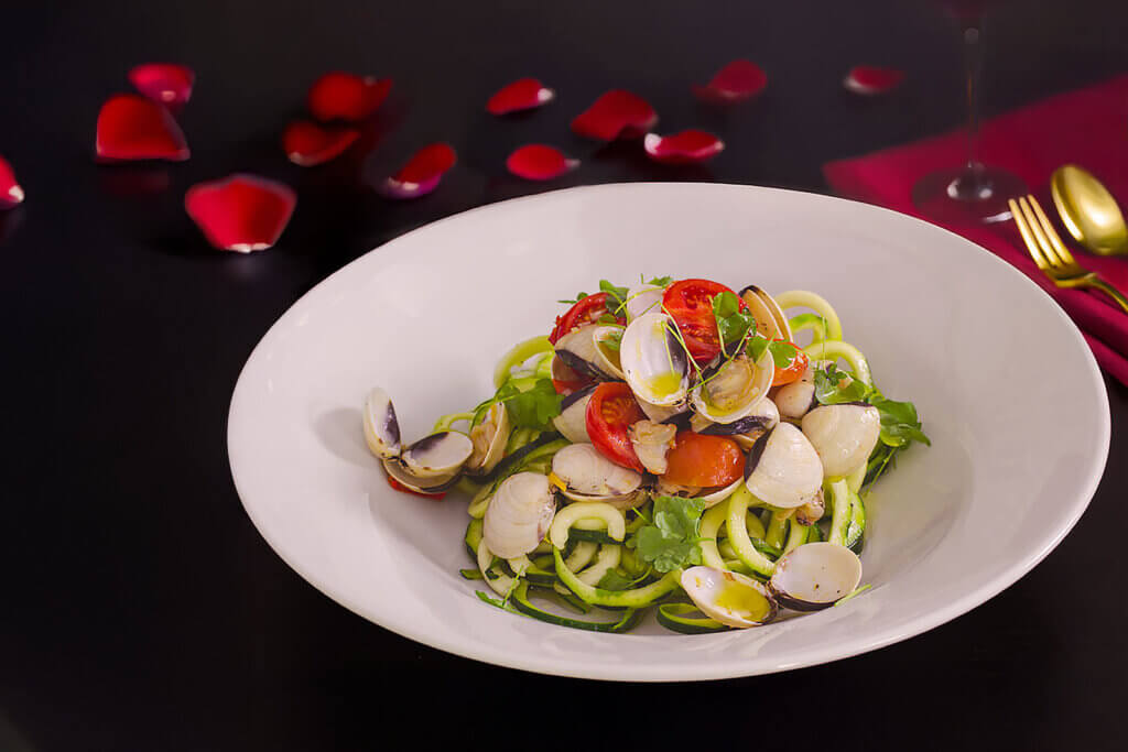 Low Carb Zudeln mit Venusmuscheln auf weißem Teller, garniert mit Cherrytomaten und Petersilie, umgeben von roten Rosenblättern und goldenem Besteck auf dunklem Hintergrund