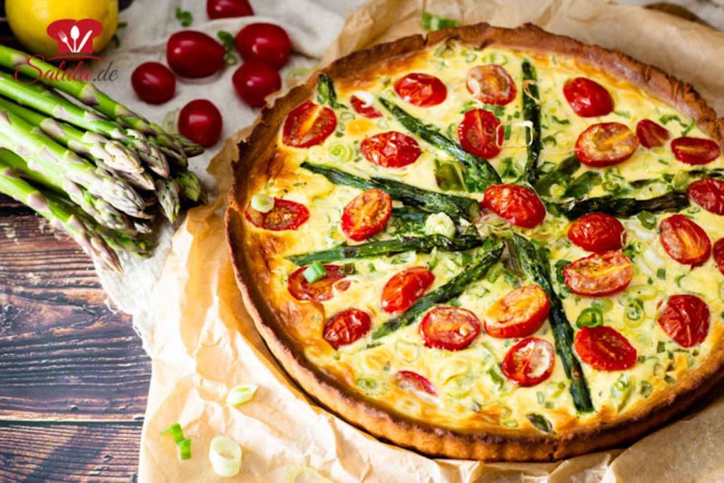 Saftige grüne Spargeltarte mit Kirschtomaten und Frühlingszwiebeln, auf einem Holzbrett mit frischem grünen Spargel und Kirschtomaten daneben