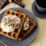 Zwei Schoko Chaffles auf einem grauen Teller mit einer Haube aus Schlagsahne und Schokostreuseln, daneben eine Tasse Kaffee.