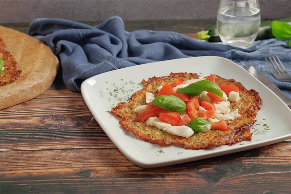 Ein Teller mit einer Low Carb Zucchini-Pizza, belegt mit Käse und Tomaten, auf einem Holztisch. Im Hintergrund ist ein Glas Wasser zu sehen.