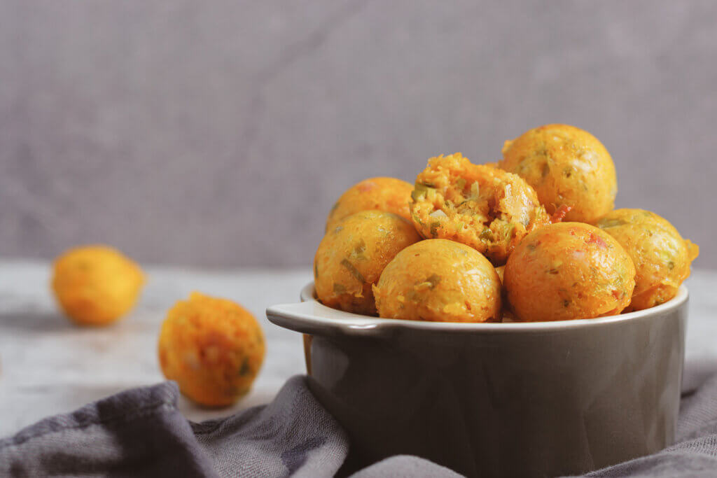 Eine Schüssel mit Low Carb Käsebällchen, die rund und orange sind. Die Schüssel ist dunkelgrau und steht auf einer hellgrauen Arbeitsplatte. Einige Käsebällchen liegen neben der Schüssel. Der Hintergrund ist hellviolett mit einer unscharfen Textur. Neben der Schüssel liegt eine graue Stoffserviette.