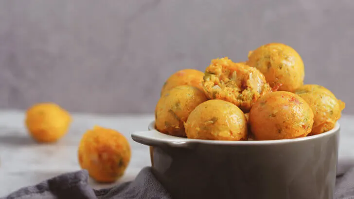 Eine Schüssel mit Low Carb Käsebällchen, die rund und orange sind. Die Schüssel ist dunkelgrau und steht auf einer hellgrauen Arbeitsplatte. Einige Käsebällchen liegen neben der Schüssel. Der Hintergrund ist hellviolett mit einer unscharfen Textur. Neben der Schüssel liegt eine graue Stoffserviette.