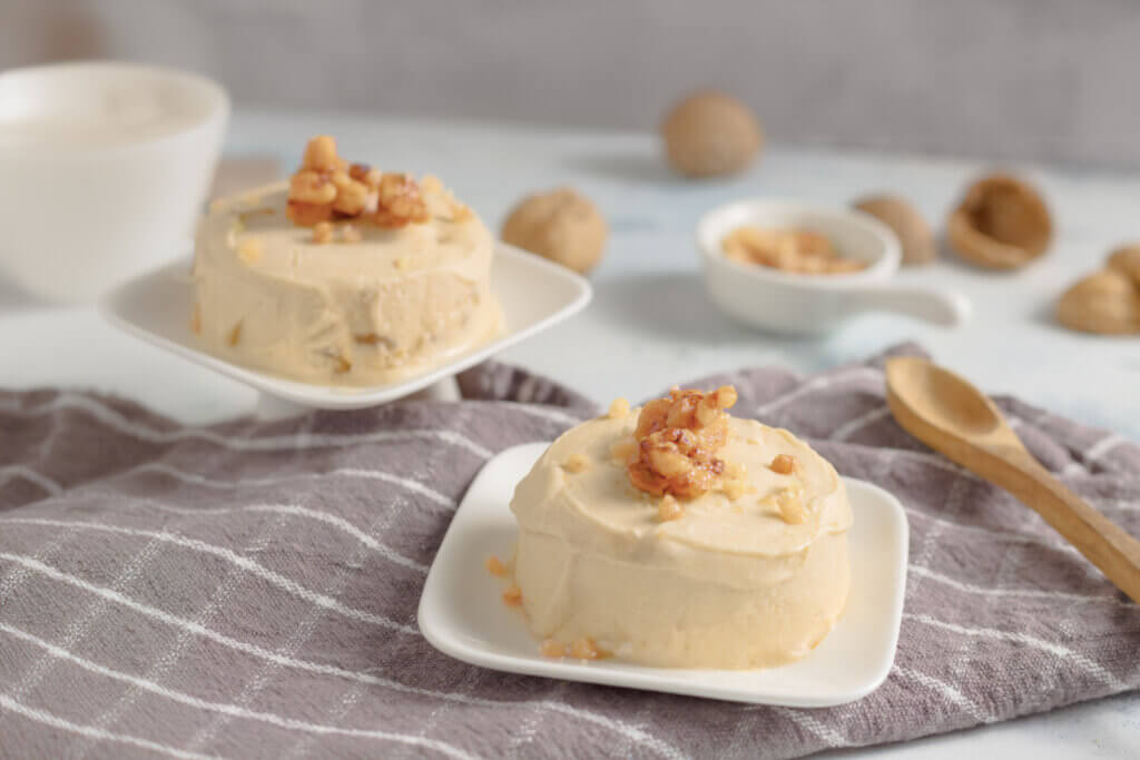 Zuckerfreies Keto-Walnusseis in zwei Portionen, gekrönt mit goldbraunem Walnusskrokant. Begleitet von einer weißen Kaffeetasse und ganzen Walnüssen im Hintergrund