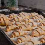 Auf einem Backblech liegen viele frisch gebackene Kanelbullar Hörnchen, die mit Puderzucker bestäubt sind. Im Hintergrund stehen grüne Weihnachtsbäume.