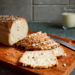Ein angeschnittenes Low Carb Kefir-Sonnenblumenkernbrot mit Sonnenblumenkernen bestreut, auf einem Holzbrett liegend, daneben ein Messer und ein Glas Milch.