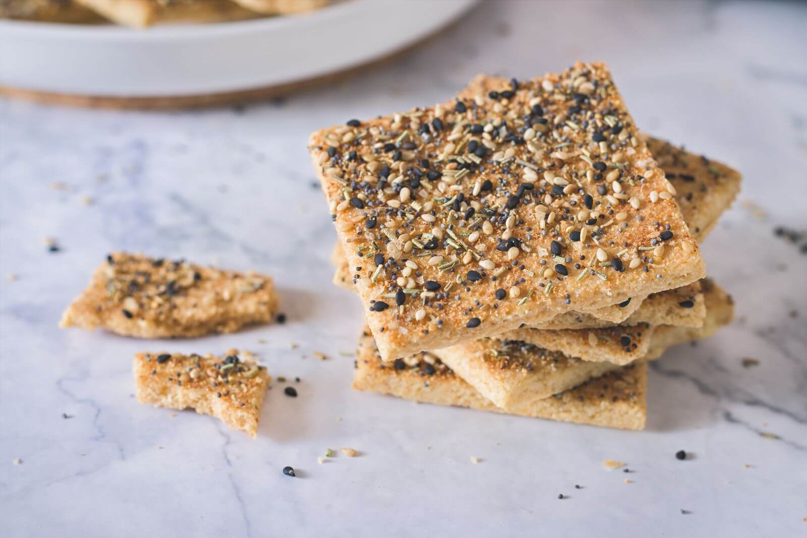 Ein Stapel goldbraun gebackener, quadratischer Keto Cracker, bestreut mit "Everything Bagel"-Gewürz (Sesam, Mohn, etc.), auf einer hellen Marmoroberfläche. Daneben liegen zwei abgebrochene, krümelige Stücke und lose Samen. Im unscharfen Hintergrund steht ein weißer Teller.