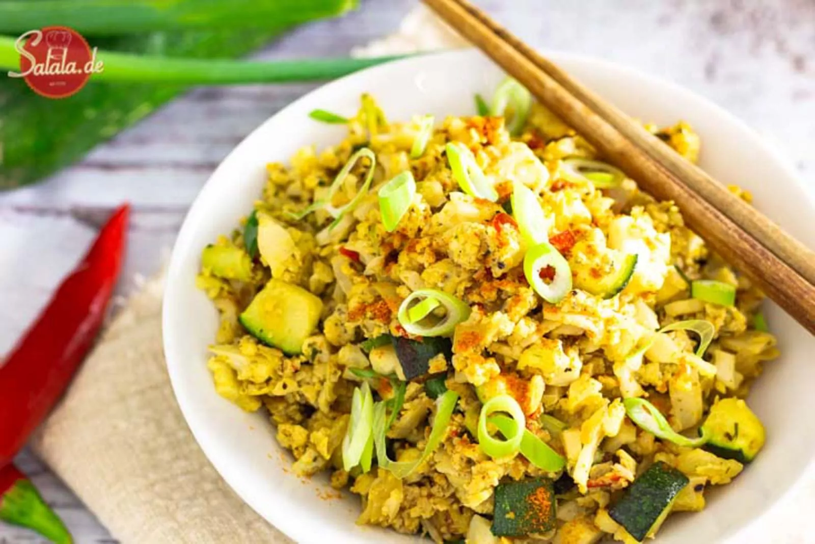 Eine Schüssel mit gebratenem Blumenkohlreis, Zucchini und Lauchzwiebeln. Zwei Holzstäbchen liegen darauf. Das Logo Salala.de ist im Hintergrund zu sehen.