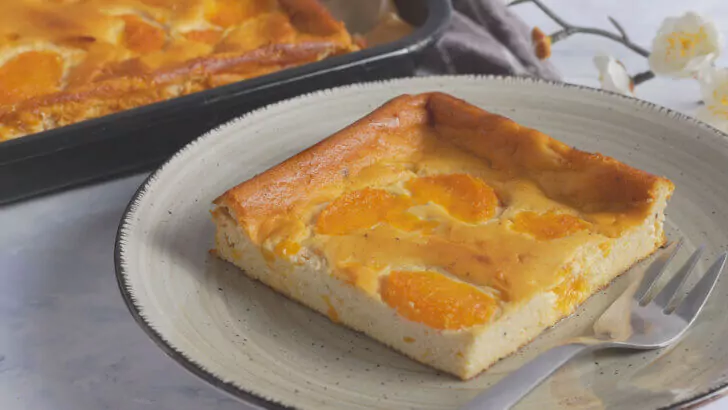 Ein Stück Mandarinenkuchen auf einem weißen Teller mit beigem Rand und einem dezenten Muster, daneben liegt eine silberne Gabel. Im Hintergrund ist ein größeres Tablett mit dem gleichen Kuchen und einige weiße Blumen zu sehen. Der Hintergrund ist eine weiße Marmor-Arbeitsplatte.