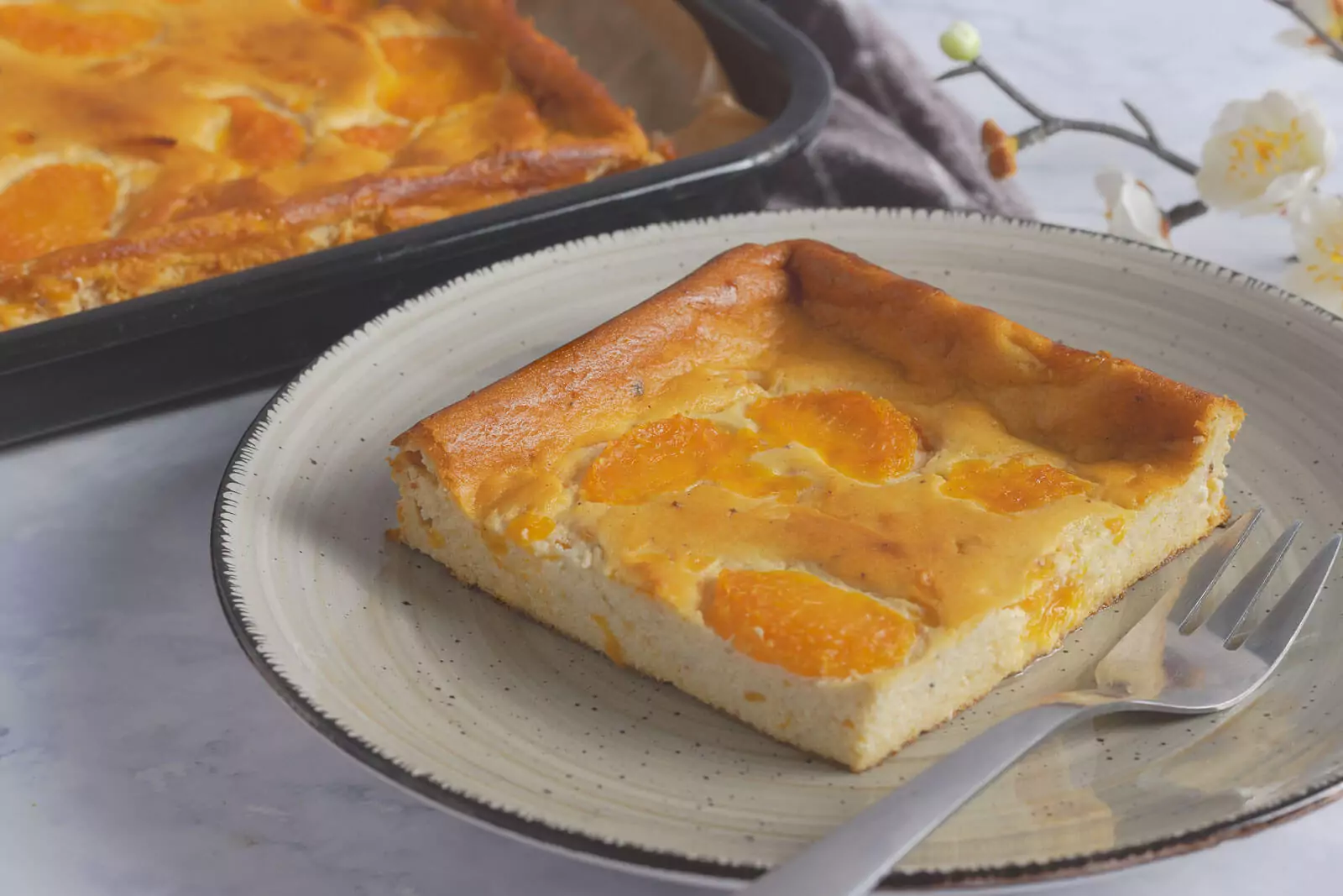 Ein Stück Mandarinenkuchen auf einem weißen Teller mit beigem Rand und einem dezenten Muster, daneben liegt eine silberne Gabel. Im Hintergrund ist ein größeres Tablett mit dem gleichen Kuchen und einige weiße Blumen zu sehen. Der Hintergrund ist eine weiße Marmor-Arbeitsplatte.