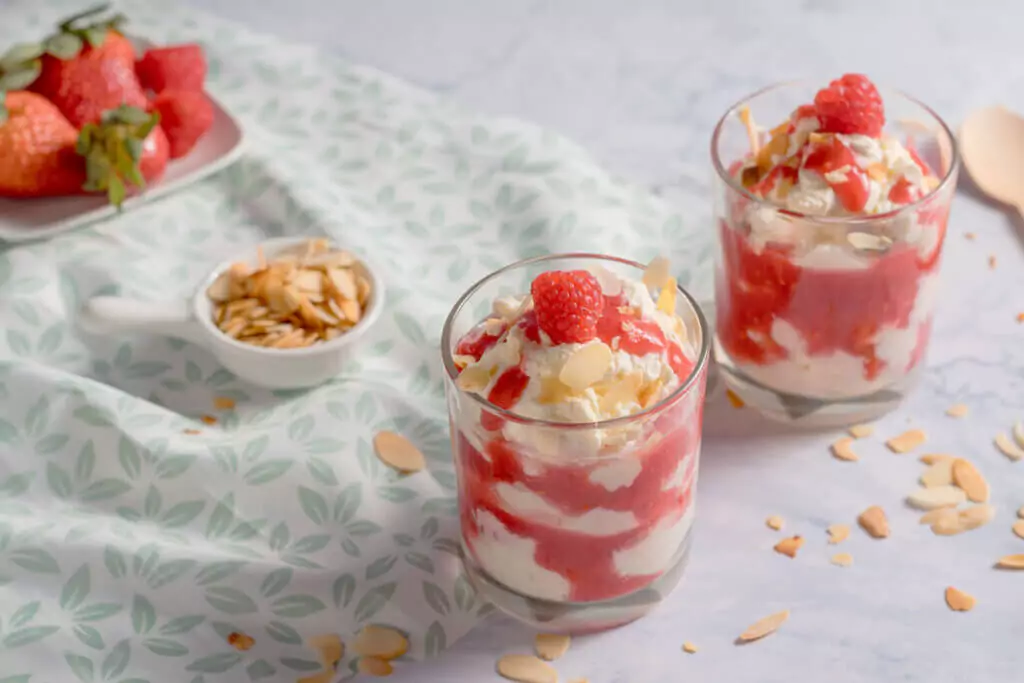 Zwei Gläser mit cremiger Cheesecake-Creme und Beerensauce, dekoriert mit Himbeeren und Mandeln auf einer gemusterten Tischdecke