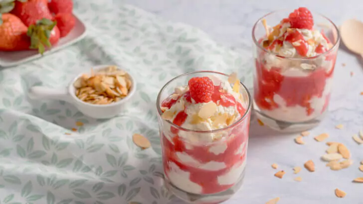 Zwei Gl&auml;ser mit cremiger Cheesecake-Creme und Beerensauce, dekoriert mit Himbeeren und Mandeln auf einer gemusterten Tischdecke