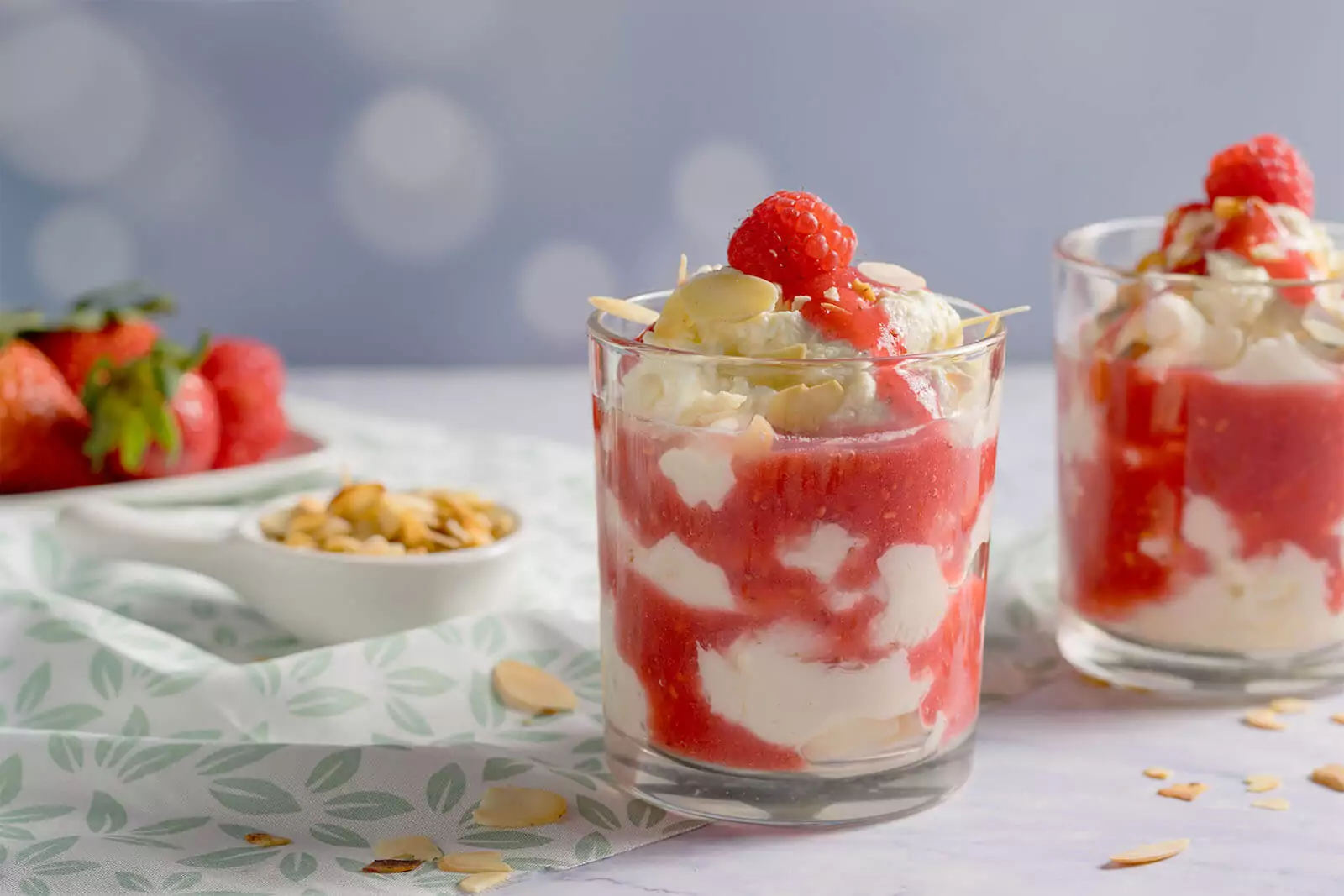 Zwei Gl&auml;ser mit cremiger Cheesecake-Creme und Beerensauce, dekoriert mit Himbeeren und Mandeln auf einer gemusterten Tischdecke.