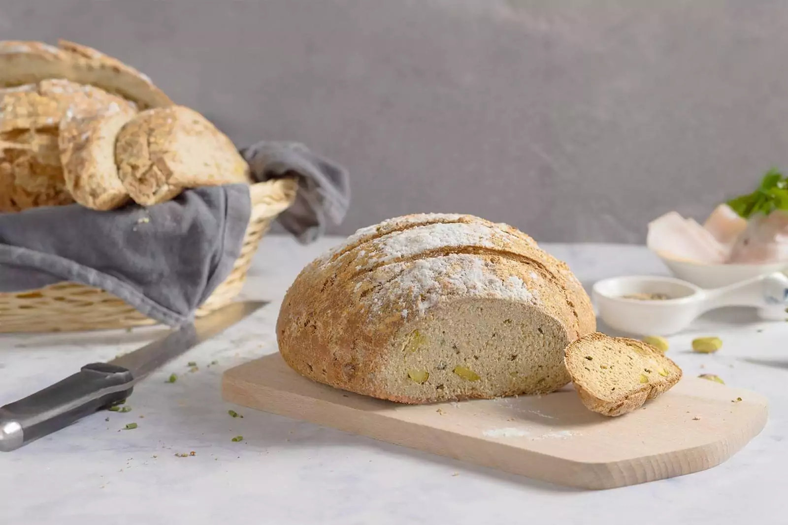 Ein rundes Laib Low Carb und glutenfreies Brot, das auf einem h&ouml;lzernen Schneidebrett liegt. Das Brot ist mit feinen K&ouml;rnern und Kr&auml;utern bestreut und sieht appetitlich aus. Im Hintergrund ist ein Brotkorb zu sehen.