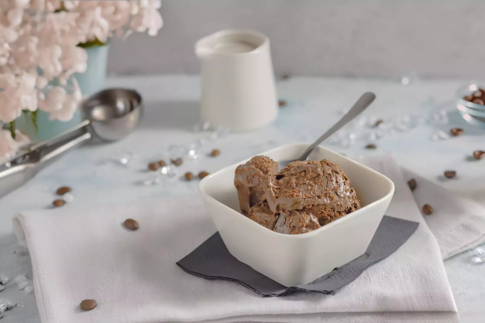 Schüssel mit Mokka-Eis, umgeben von Kaffeebohnen und Eisdekoration auf einem weißen Tuch. Im Hintergrund eine Eisportionierer, eine Vase mit rosa Blumen und ein weißer Krug.