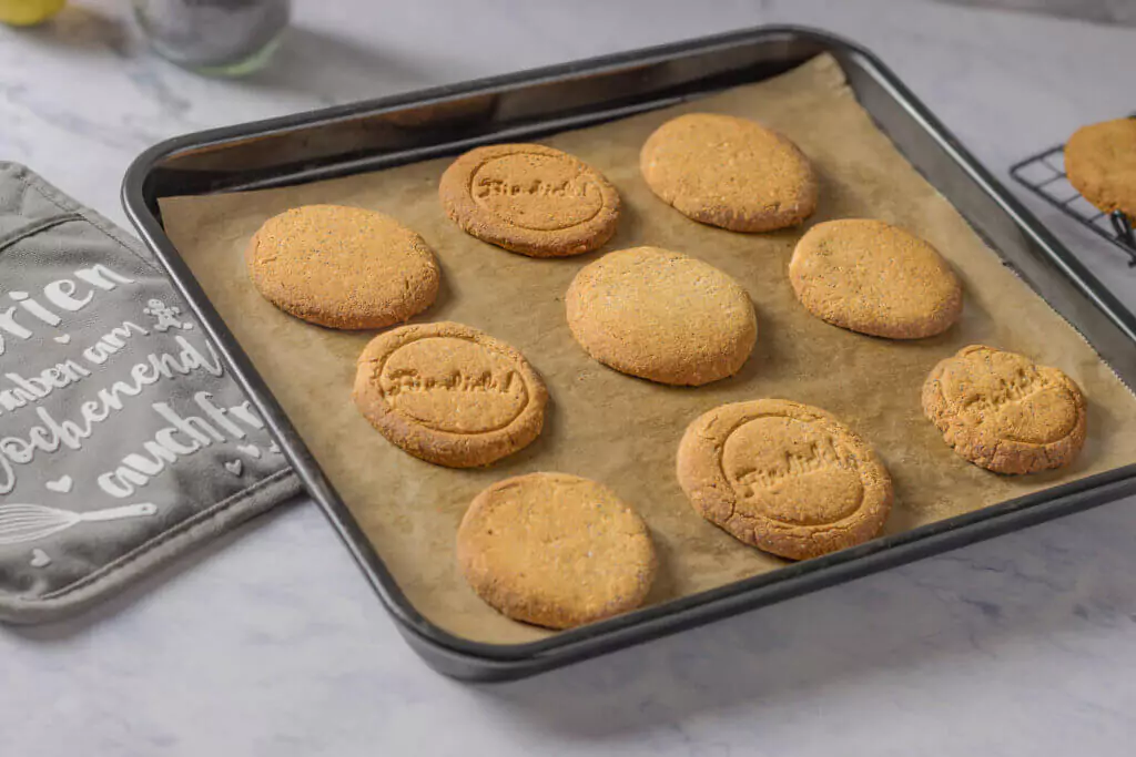 Auf einem Backblech liegen mehrere Zitrone-Mohn-Kekse ohne Zucker, die auf Backpapier gebacken wurden.