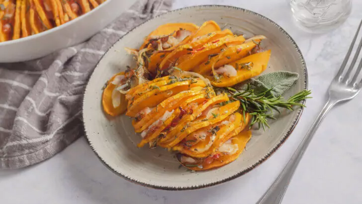 Keto-freundliches K&uuml;rbisgratin auf wei&szlig;em Teller, dekoriert mit Rosmarin. Sichtbar sind auch eine Gabel, ein Wasserglas und eine Stoffserviette.