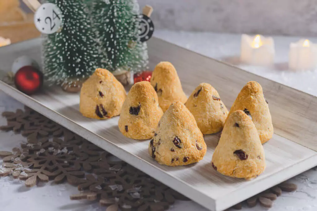 Kohlenhydratarme Cranberry-Tannenbäume aus Kokosteig auf einem Holztablett, dekoriert mit Weihnachtsaccessoires wie Kugeln, kleinen Tannenbäumen und Kerzen.